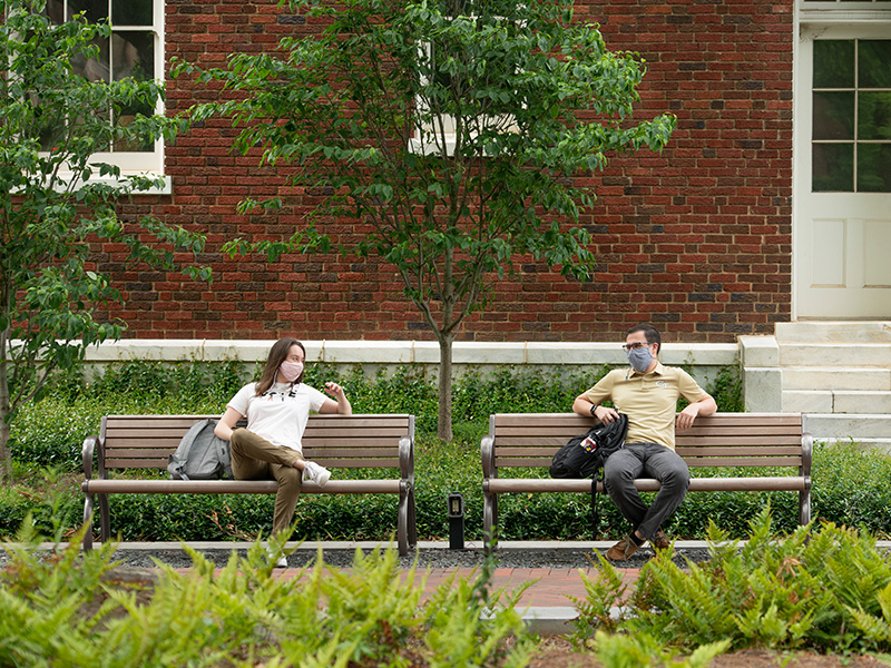Two students on separate benches, wearing masks, and talking to one another