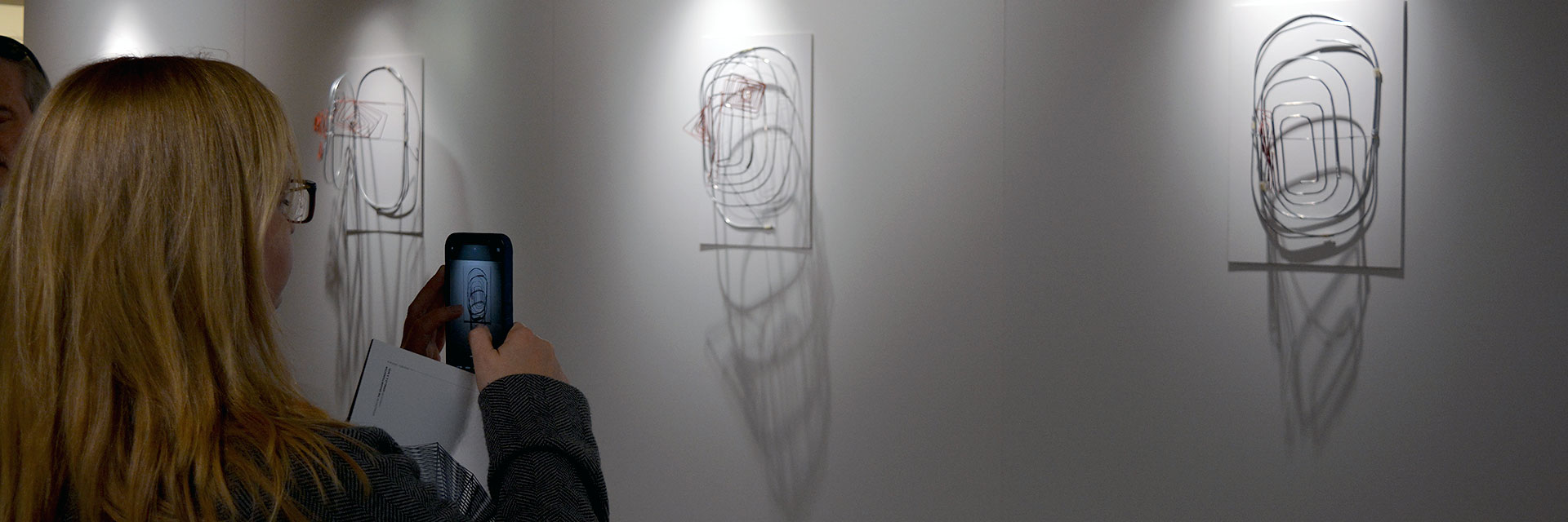 A visitor to Vernelle Noel's wire-bending exhibit takes a photograph of Carnival Masks mounted on a wall.