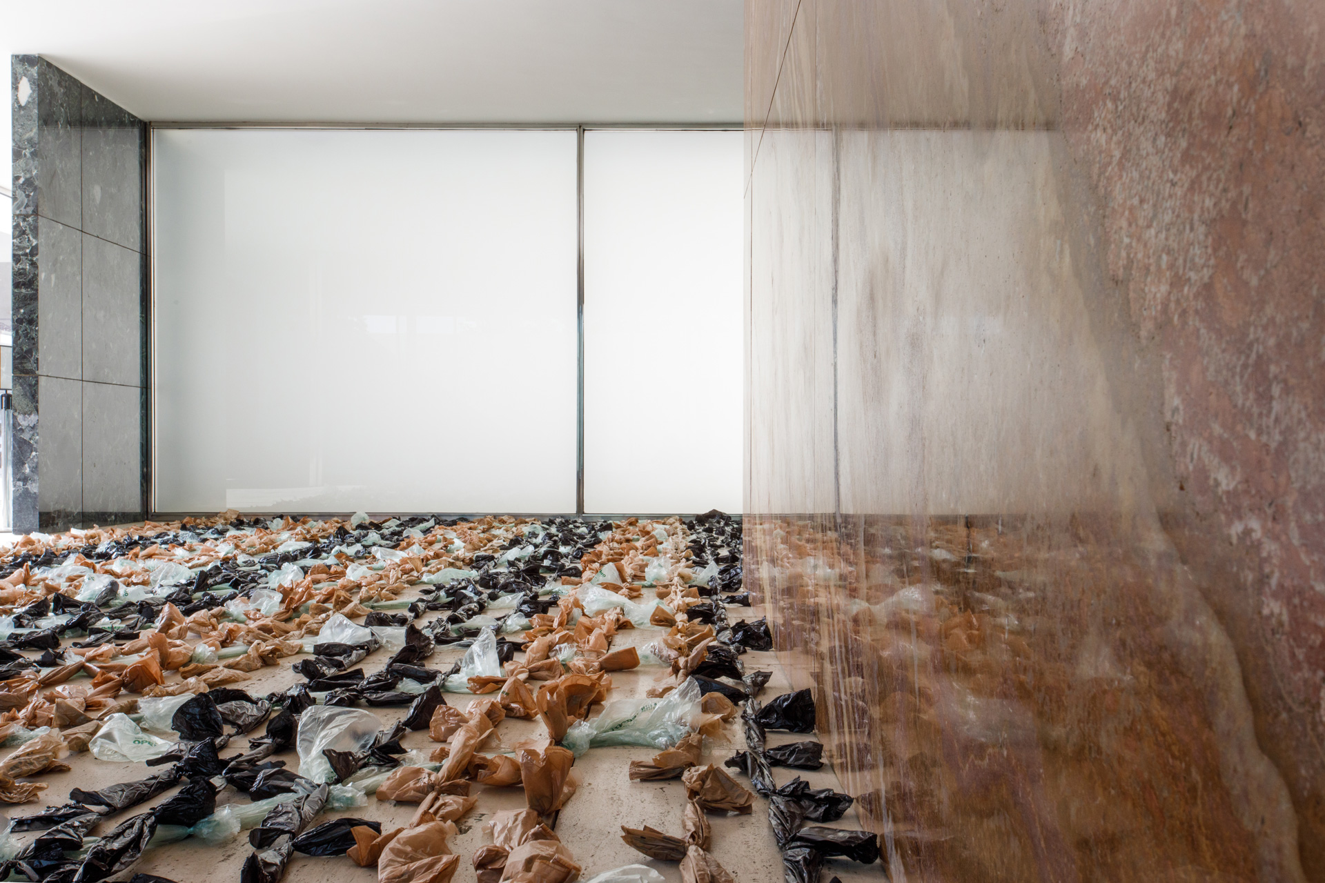 Interior shot of Mies van der Rohe Pavilion in Barcelona. A red onyx wall stretches away from the viewer, filling the right half of the shot. The floor is covered by strands of disposable shopping bags.